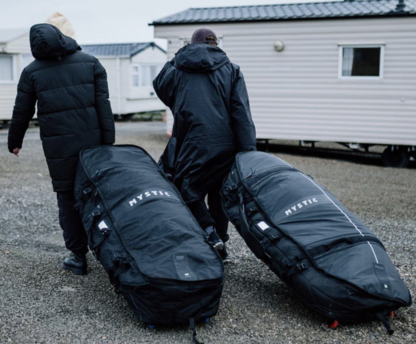 Two travellers in winter coats wheeling large Mystic board bags across gravel past a static caravan