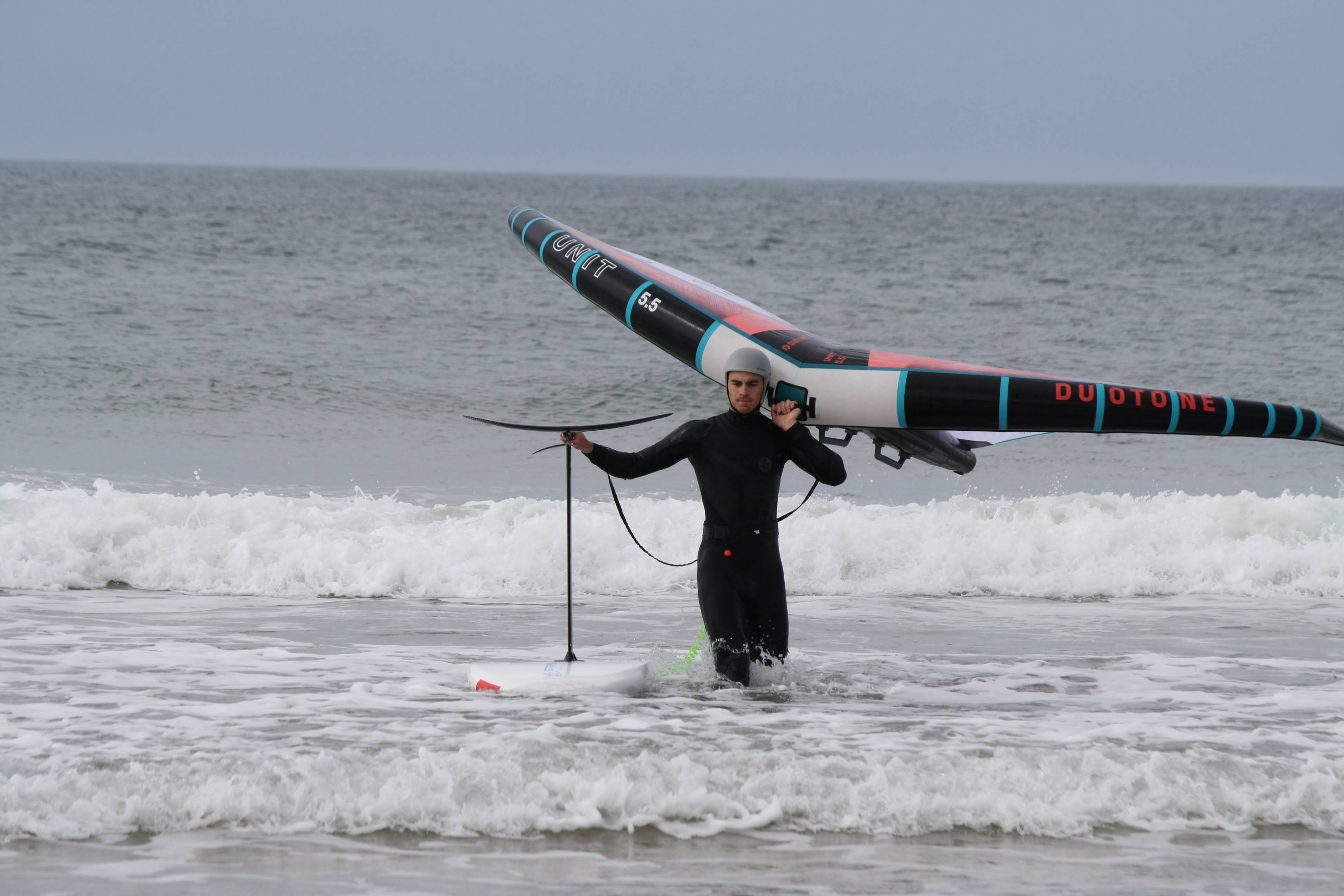 Wing foiler in a black wetsuit standing in the shallows holding his wing and board
