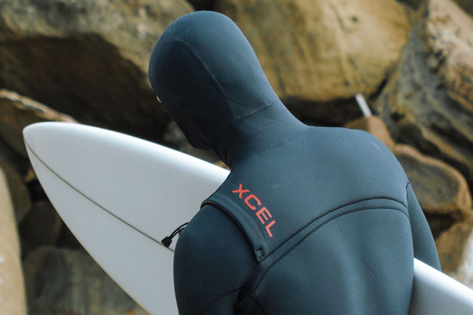 Close-up of a surfer's back in a hooded dark teal Xcel wetsuit, carrying a white surfboard past rocks