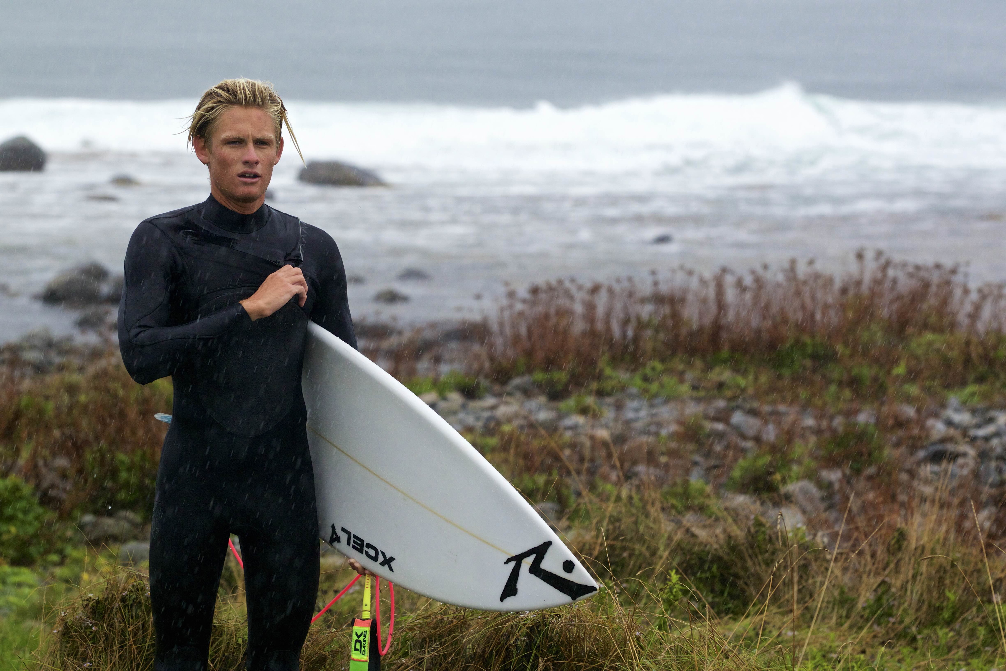 Surfer in a black wetsuit standing in coastal grass with a white surfboard under his arm