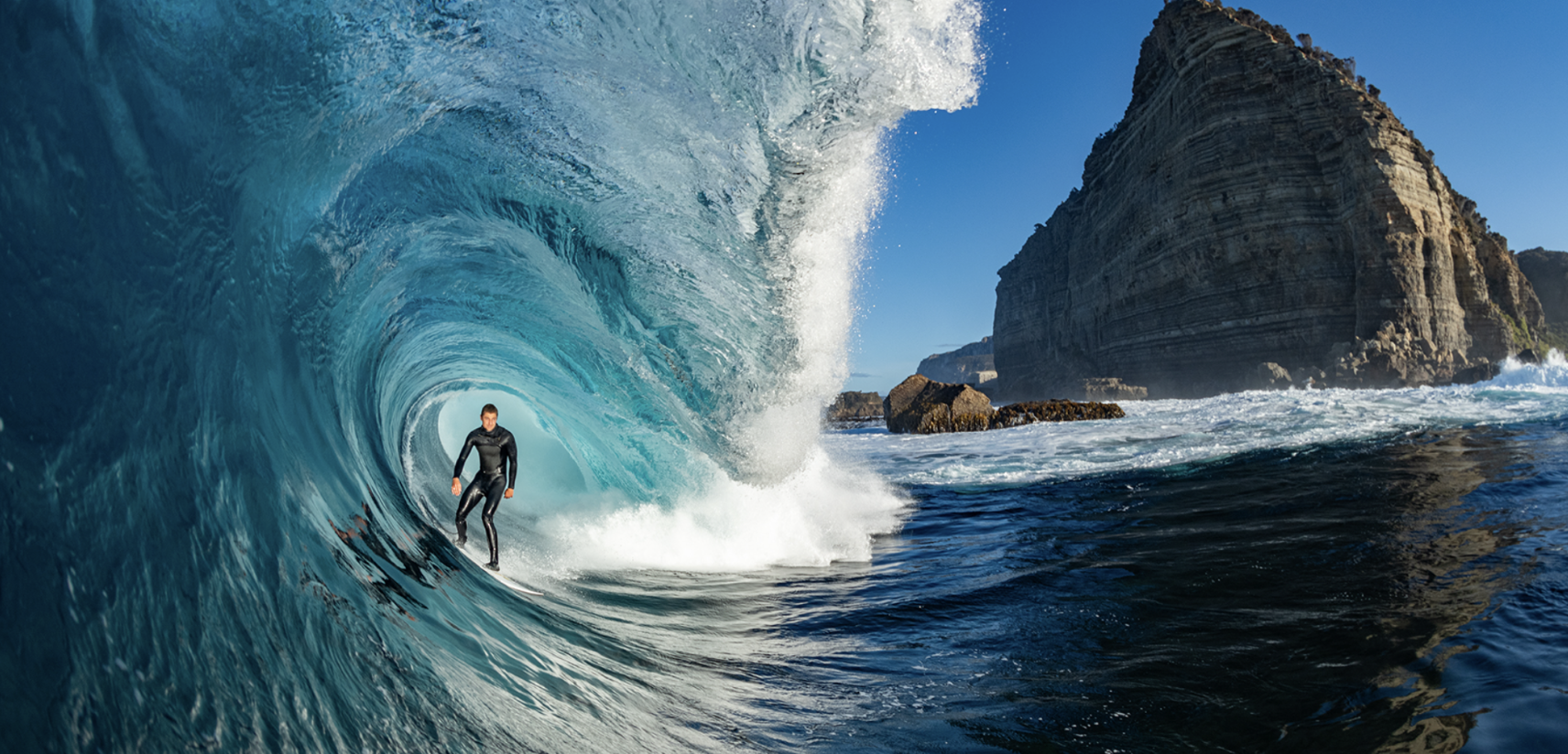 Surfer riding deep inside a large barrelling wave beneath a towering cliff