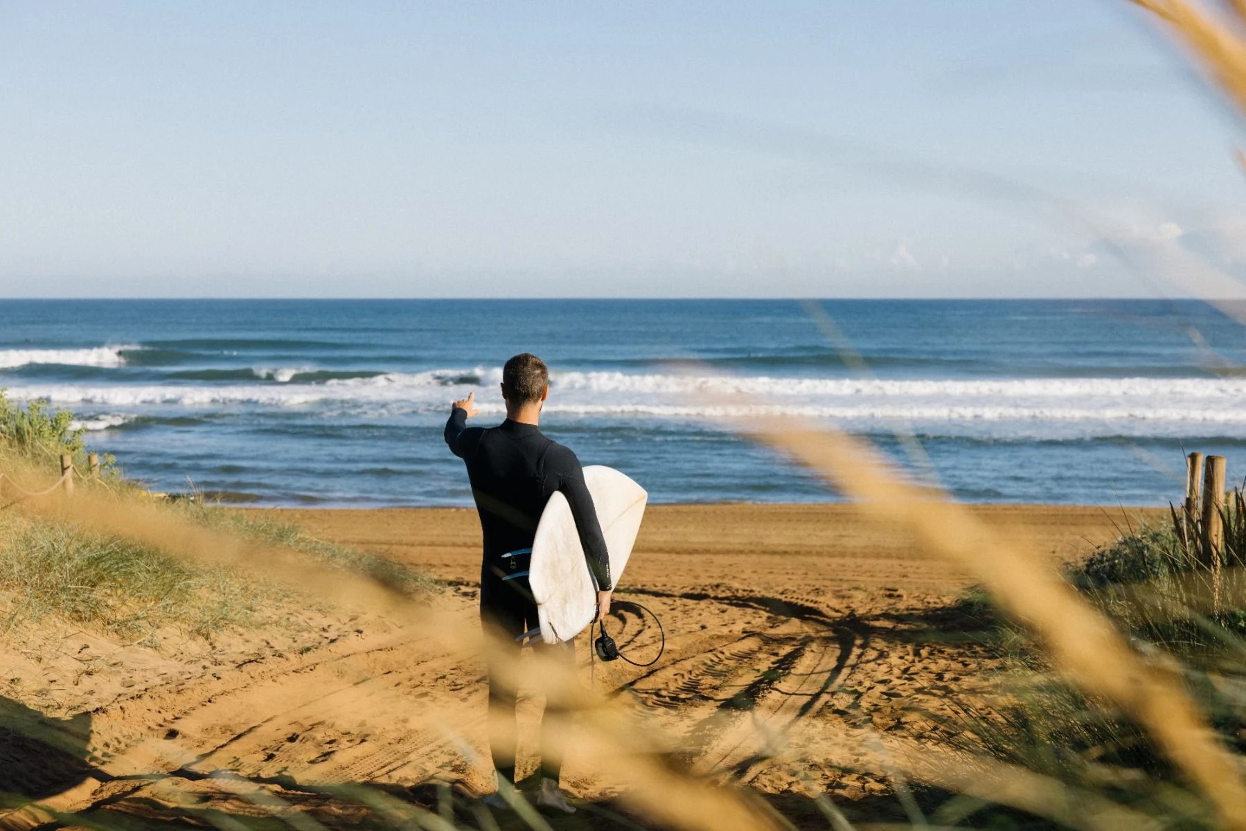 Surfista in muta nera che scende un sentiero sabbioso tra le dune verso la spiaggia con la tavola sotto il braccio