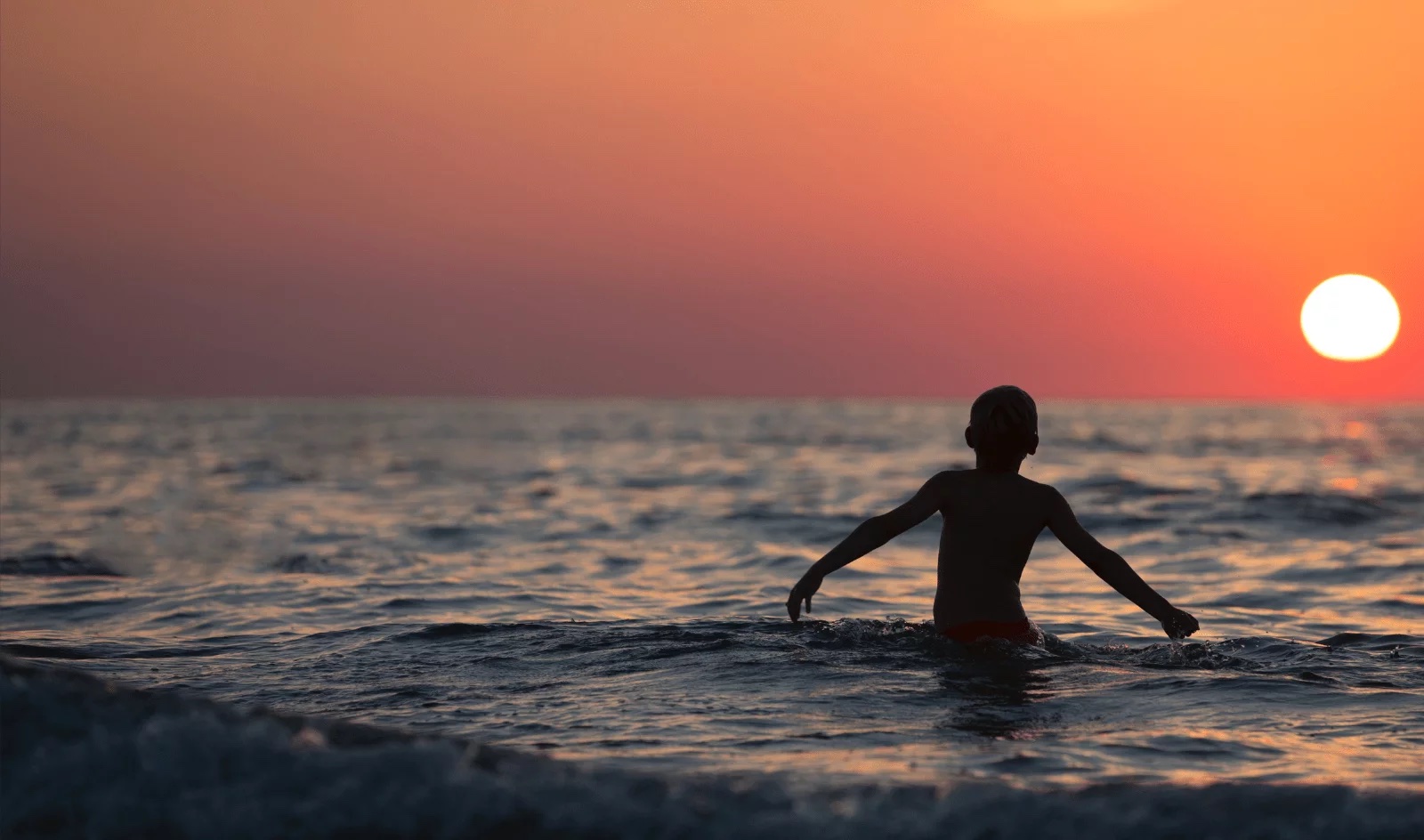 Silhouetted child standing waist deep in the sea as the sun sets over the horizon