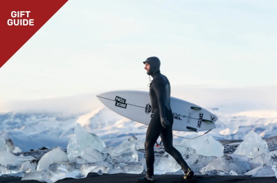 Surfista in muta invernale con cappuccio che porta la tavola tra le dune
