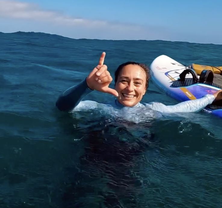 Windsurfer in the water beside her rig, grinning and throwing a shaka to the camera