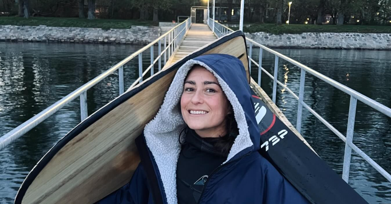 Smiling woman in a hooded jacket sitting in a small boat with her board, cool grey water behind