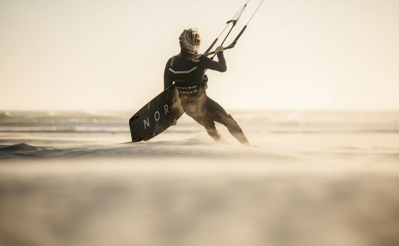 Kitesurfer in a harness carrying a board across a windswept beach in low evening light
