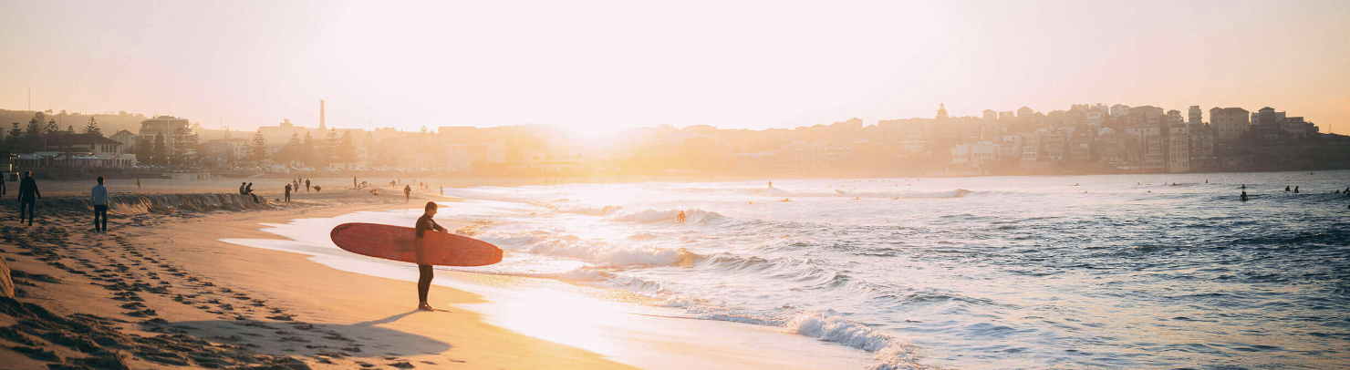 Surfer carrying a board along a wide beach at sunrise with a town on the headland behind