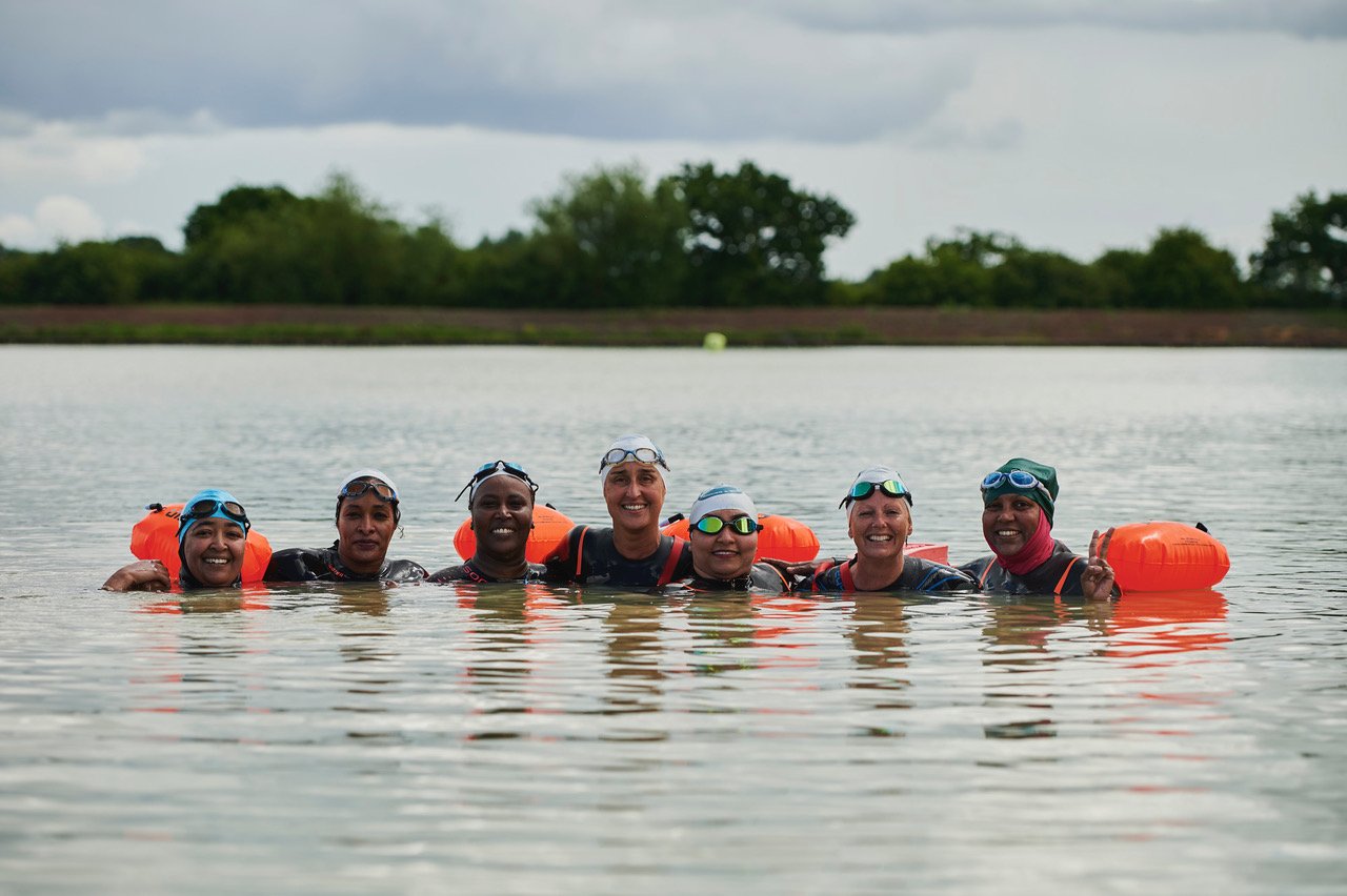 Group of open-water swimmers in caps and goggles standing chest-deep in a lake with orange tow floats